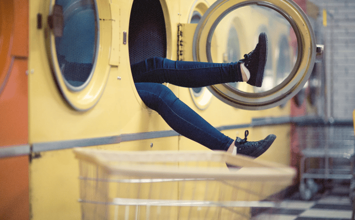 Des jambes de femmes sortent d'une machine à laver ouverte, dans un lavomatic